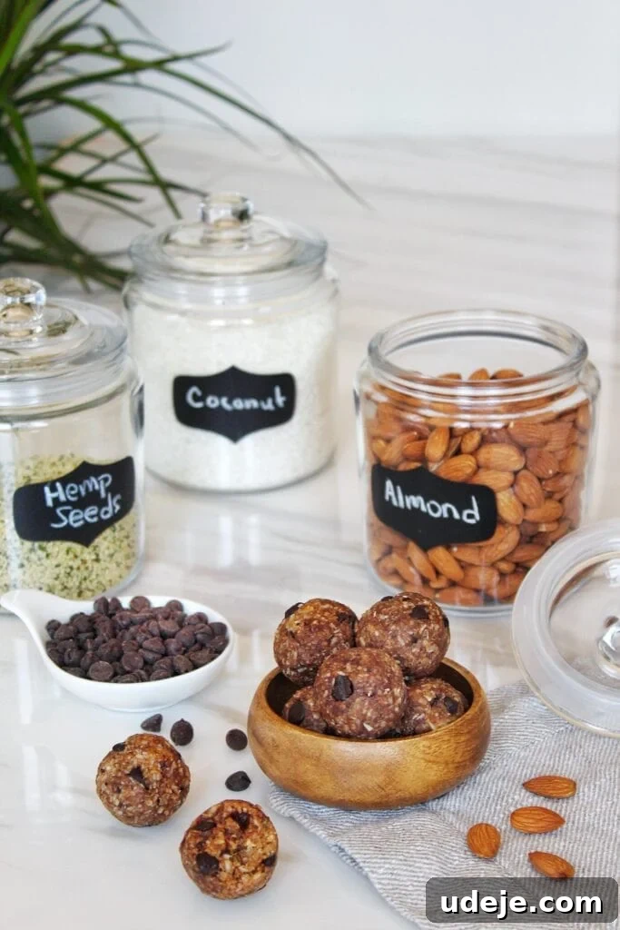 Dateless Energy Bites 2 A bowl of date-less energy bites sits on a linen cloth beside a clear jar of raw almonds. In the background, jars of shredded coconut and hemp seeds, along with a small dish of dark chocolate chips, are arranged on a white marble countertop, creating a clean and inviting scene. A green plant adds a touch of freshness.