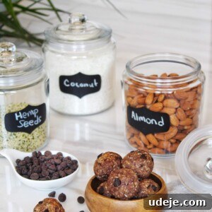 Dateless Energy Bites 3 A wooden bowl overflowing with homemade date-less energy bites, artfully arranged on a soft linen cloth. Surrounding the bowl are various ingredients: a rustic glass jar filled with raw almonds, another jar showcasing delicate coconut flakes, a third containing nutrient-rich hemp seeds, and a small, inviting dish of dark chocolate chips. A few loose whole almonds are scattered on the pristine marble countertop, completing this wholesome and appetizing display.