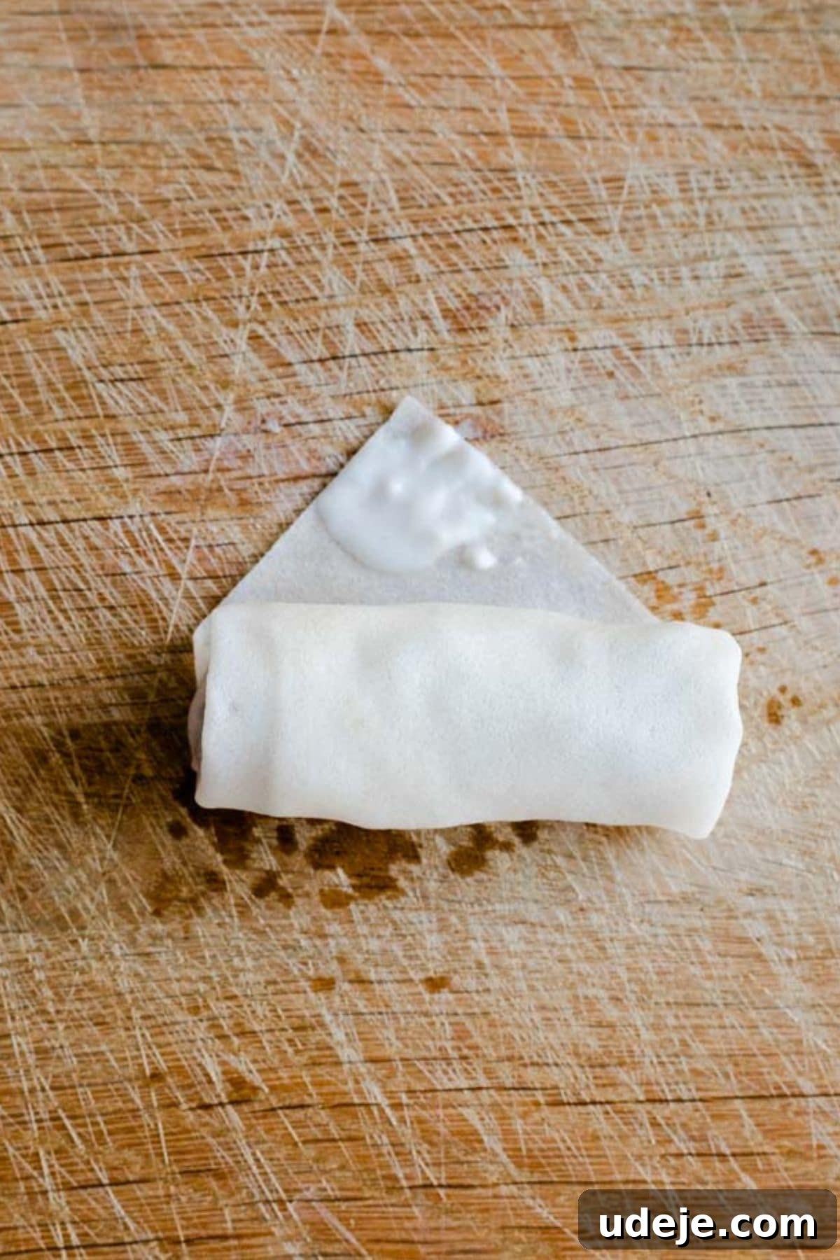 Your Guide to Authentic Homemade Sambousek 17 a square spring roll wrapper on a wooden board, with ground beef filling inside, being rolled into a cylinder shape