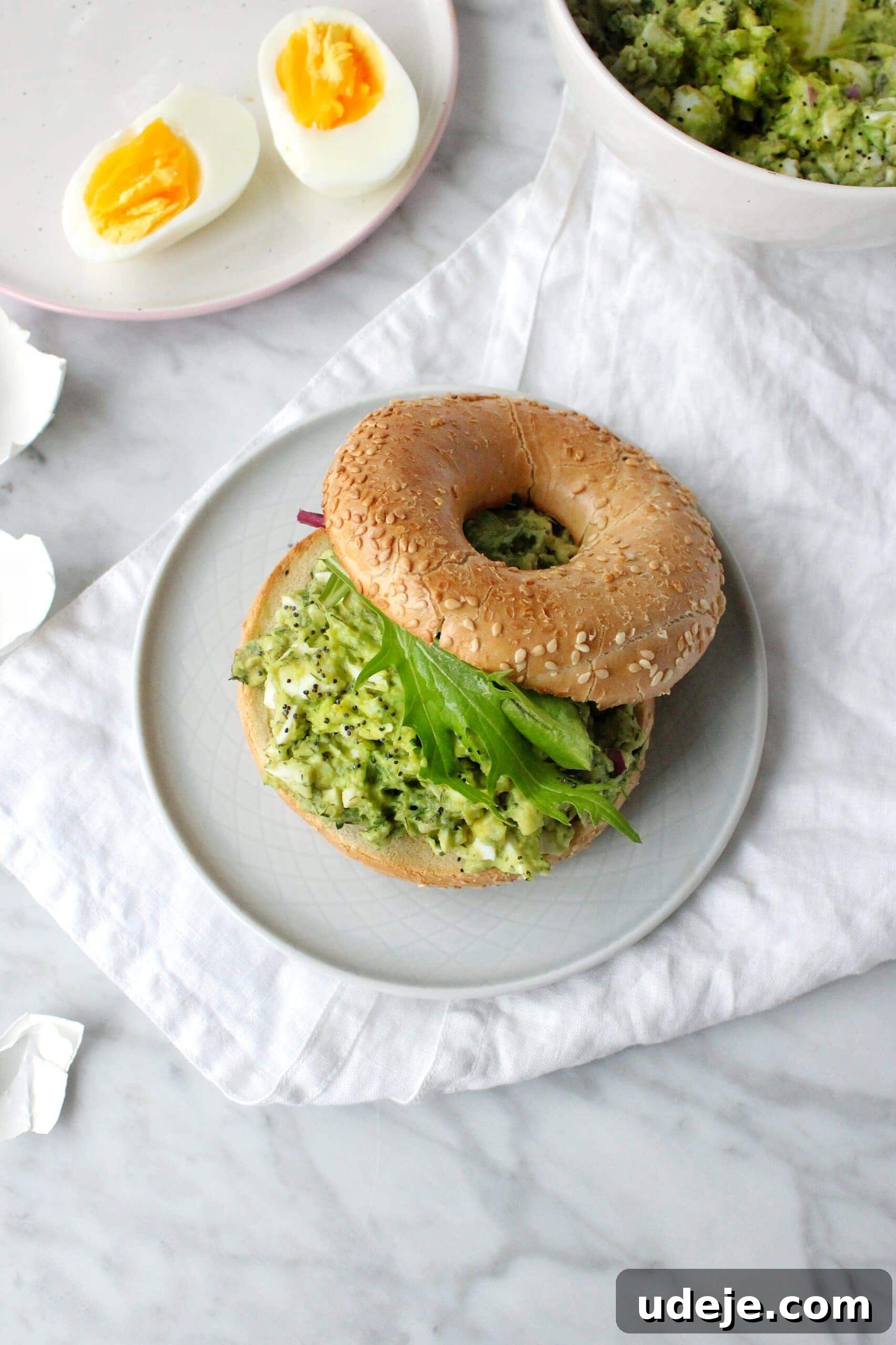 Avocado Egg Salad Supreme 2 A sesame seed bagel sandwich filled with green avocado salad and leafy greens sits on a white plate. Nearby, a plate holds two halves of a soft-boiled egg, and a bowl with more salad is visible on a marble surface.