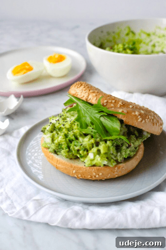 Avocado Egg Salad Supreme 3 A sesame seed bagel sandwich filled with mashed avocado and leafy greens sits on a plate. In the background, a bowl of mashed avocado and a plate with two halved soft-boiled eggs are visible.