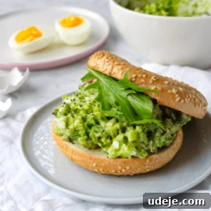 A sesame seed bagel sandwich filled with mashed avocado and leafy greens sits on a plate. In the background, a bowl of mashed avocado and a plate with two halved soft-boiled eggs are visible.