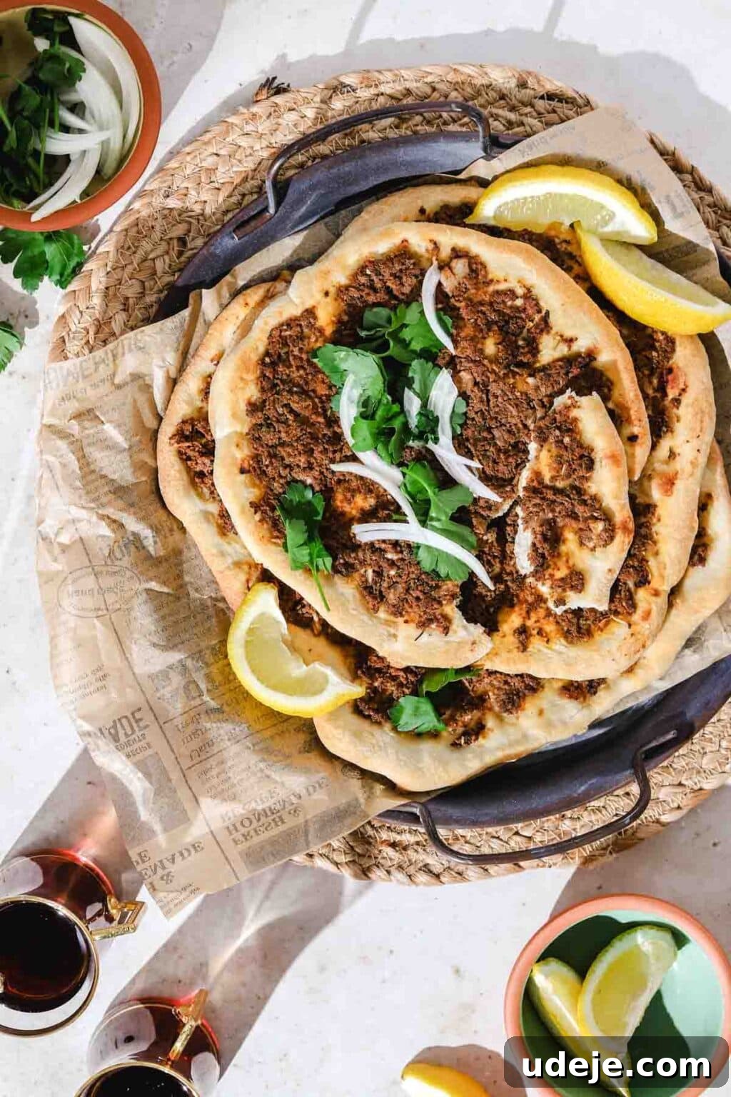Lahm Bi Ajeen Savory Middle Eastern Flatbread 2 Lahm Bi Ajeen piled on a dark colored tray with parchment, garnished with lemon wedges, parsley and onion slices on a light colored background with a bowl of sliced onions and parsley in a bowl in the corner, lemon wedges in a bowl in the bottom corner and 2 cups of tea in the corner