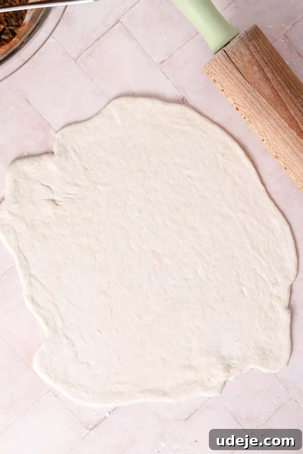 Lahm Bi Ajeen Savory Middle Eastern Flatbread 12 rolled out dough on a light pink tiled background with a wooden rolling pin in the corner and meat mixture in a glass bowl in the other top corner