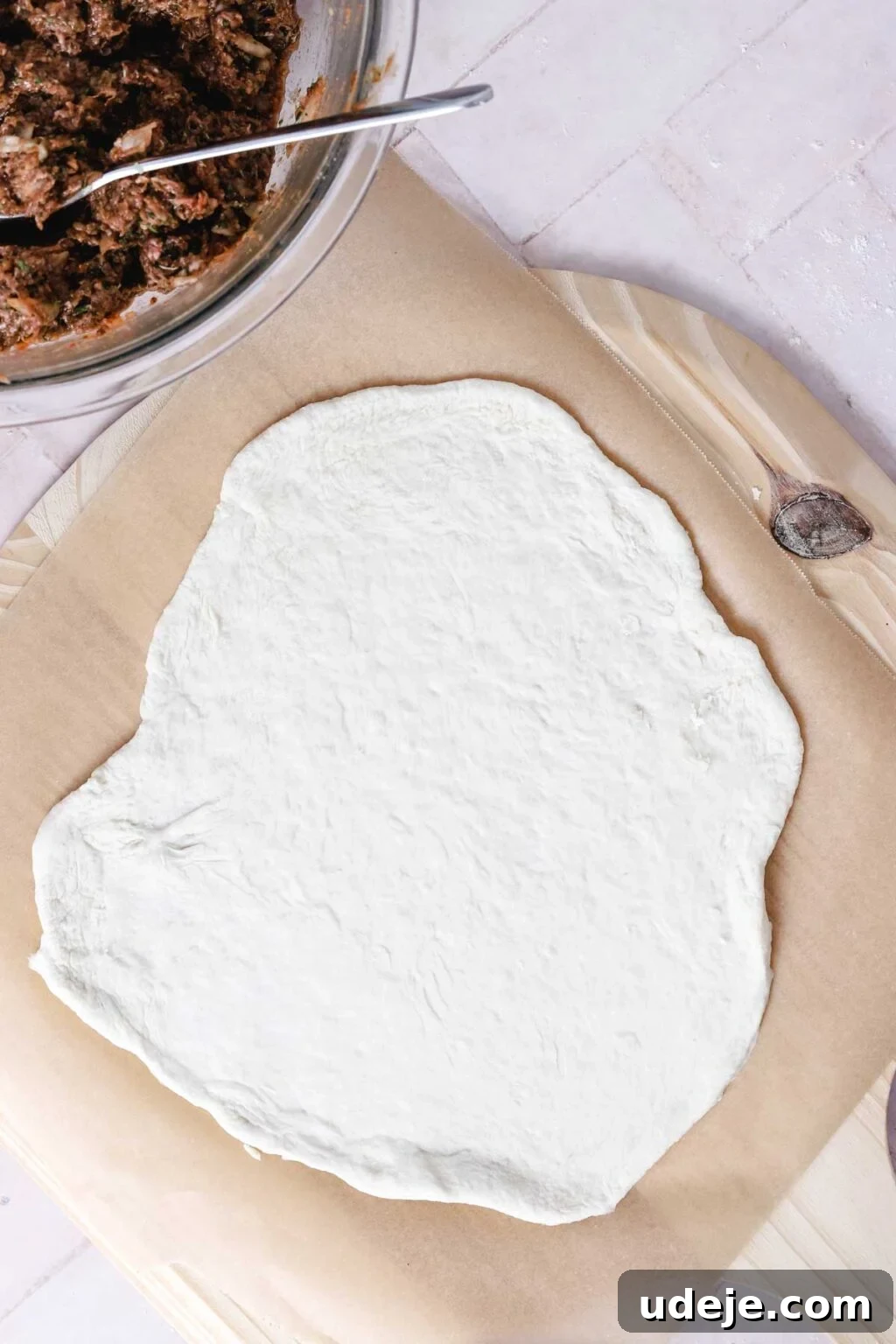 Lahm Bi Ajeen Savory Middle Eastern Flatbread 13 rolled out dough on a parchment on a wooden board on a light pink tiled background with a glass bowl with meat mixture in the corner