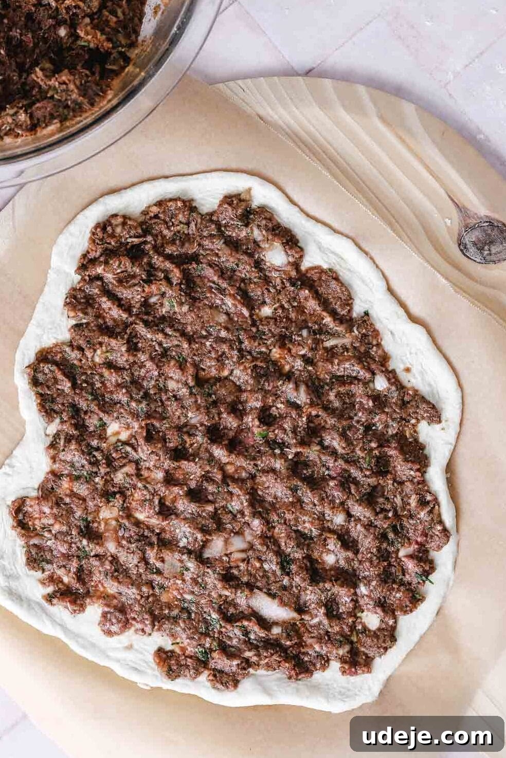 Lahm Bi Ajeen Savory Middle Eastern Flatbread 14 meat mixture spread on rolled out dough on parchment on a wooden board with a glass bowl with the meat mixture in a corner with a spoon