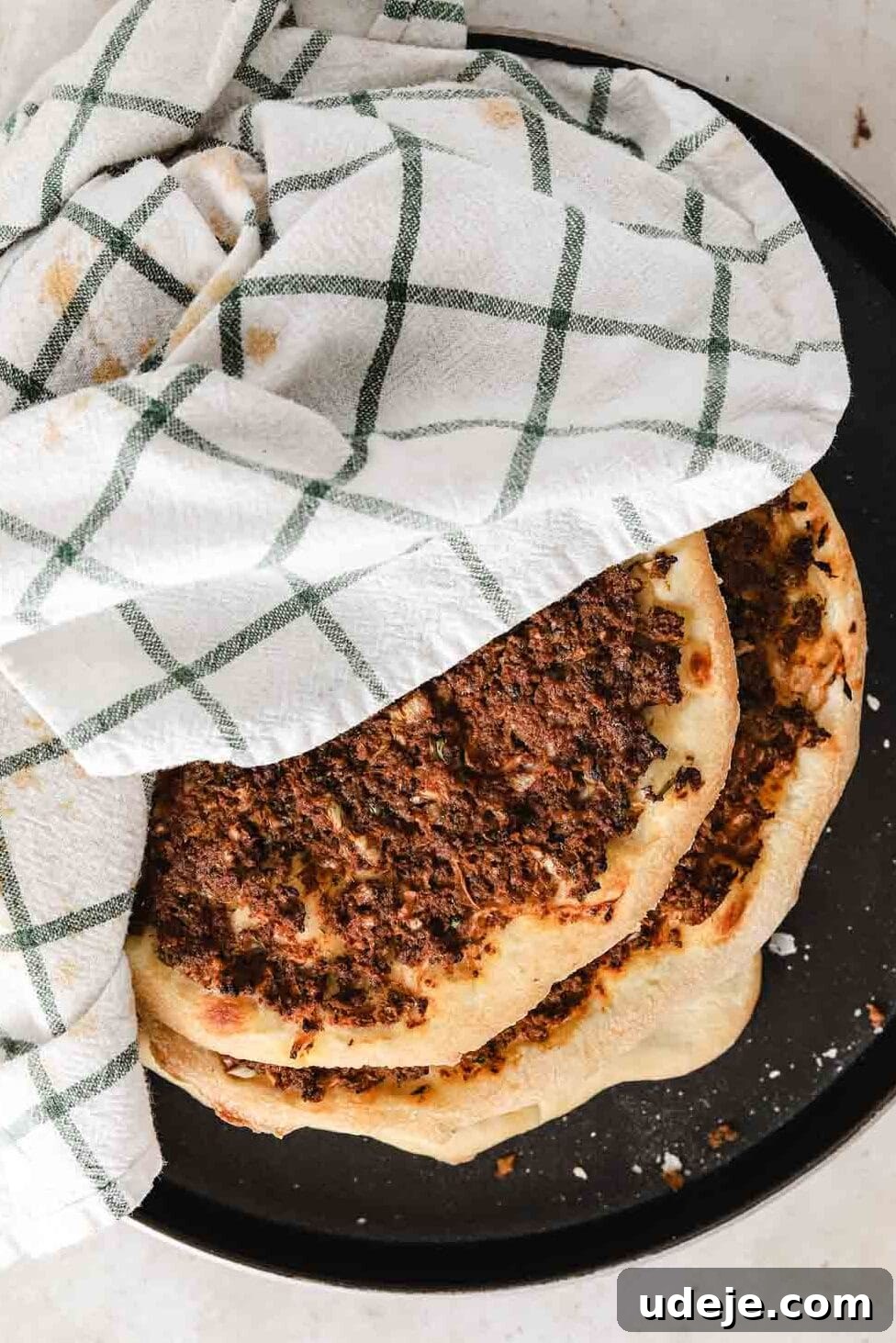 Lahm Bi Ajeen Savory Middle Eastern Flatbread 16 Lahm Bi Ajeen in a pile on a dark coloured plate with a green and white tea towel on a light coloured background