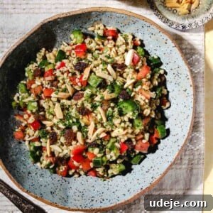 A bowl of colorful bulgur salad with diced vegetables, herbs, raisins, and slivered almonds sits on a patterned cloth next to a small dish of extra almonds and a glass of drink.