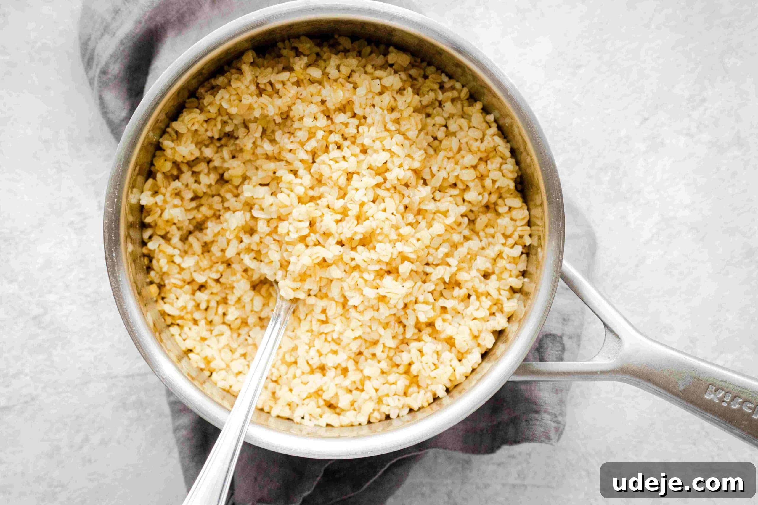 Crisp Bulgur Salad: Effortless & Delicious 4 A pot filled with cooked bulgur wheat, perfect for making Bulgur Salad, with a metal spoon resting inside, placed on a gray kitchen towel on a light surface.