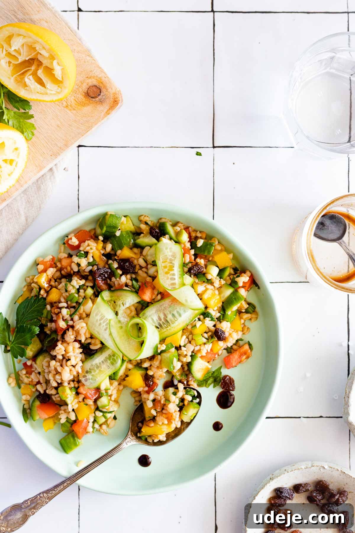 Crisp Bulgur Salad: Effortless & Delicious 6 A plate of colorful bulgur salad with cucumber ribbons, herbs, diced vegetables, and raisins sits on a tiled table next to a glass of water, a lemon half, and a small dish of dressing with a spoon.