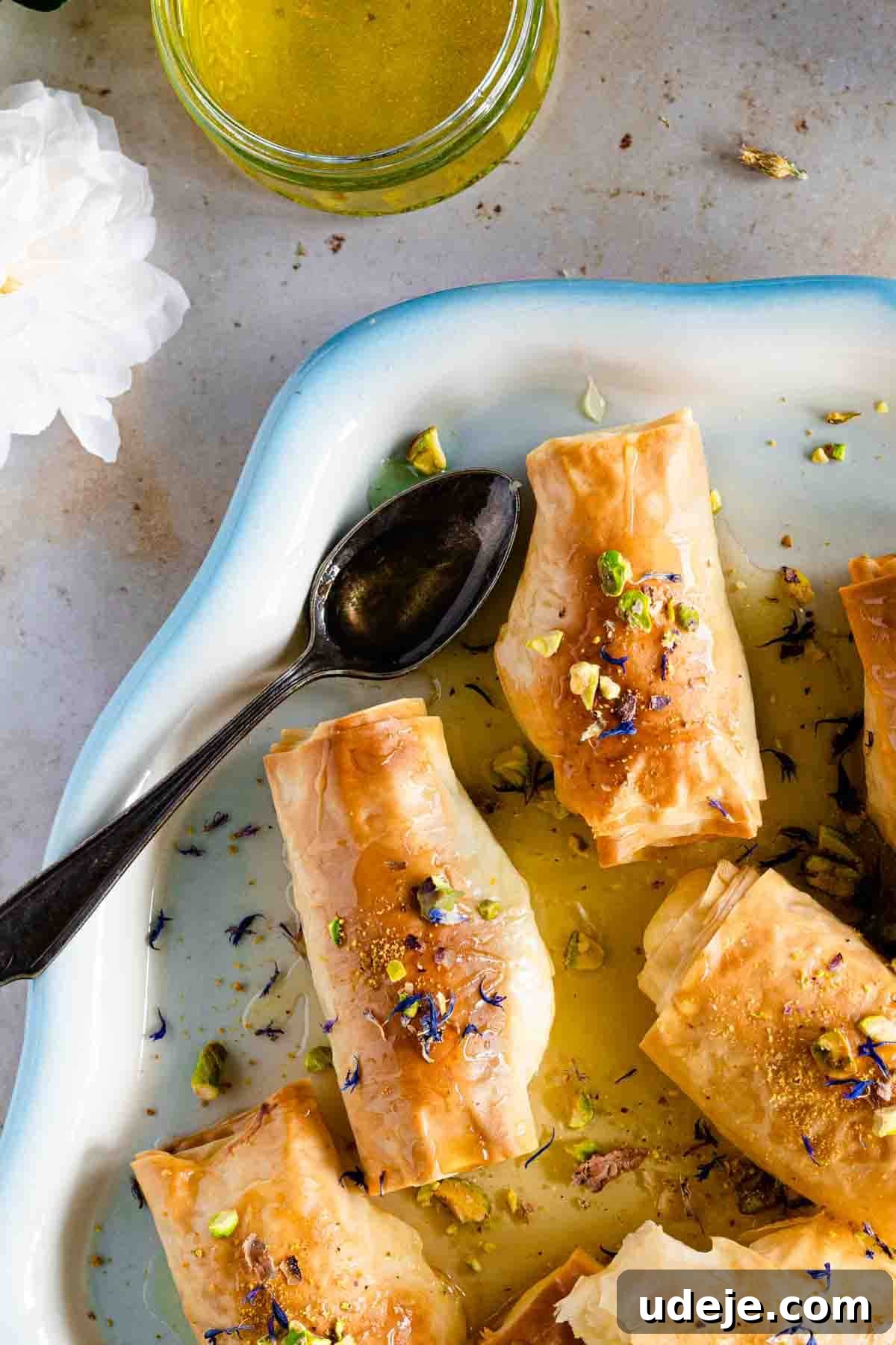 Homemade Znoud El Sit (Lady Fingers) - Crispy and Creamy Crunchy rolled phyllo dough pastries, Znoud El Sit, are elegantly presented on a white plate with a serving spoon, generously drizzled with shimmering sugar syrup and garnished with a sprinkle of vibrant green pistachios, embodying a perfect Middle Eastern dessert.
