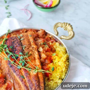 A silver bowl with two ornate handles holds Arabic Fish—spiced grilled fillets served over yellow rice with a tomato-based sauce and fresh herbs. In the background are lemon wedges, red onion slices, and fresh greens.