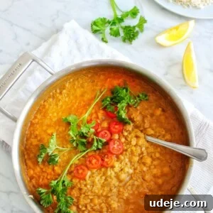 A close-up of a bubbling pot of red lentil dal garnished with fresh parsley and sliced red chili, resting on a white cloth next to lemon wedges and a plate of rice on a marble surface.