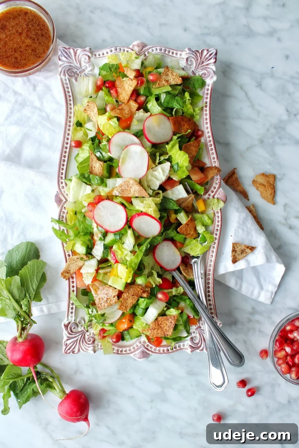 Signature Restaurant Fattoush 2 A rectangular platter of fresh Fattoush salad with lettuce, radish slices, cherry tomatoes, pita chips, and green onions. Two forks rest in the salad. Nearby are a drink, whole radishes, extra pita chips, and scattered pomegranate seeds.