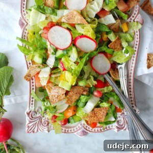 A vibrant Fattoush salad with sliced radishes, lettuce, chopped vegetables, pita chips, and pomegranate seeds is served on a decorative platter with two serving utensils. Radishes and greens are arranged beside the platter.