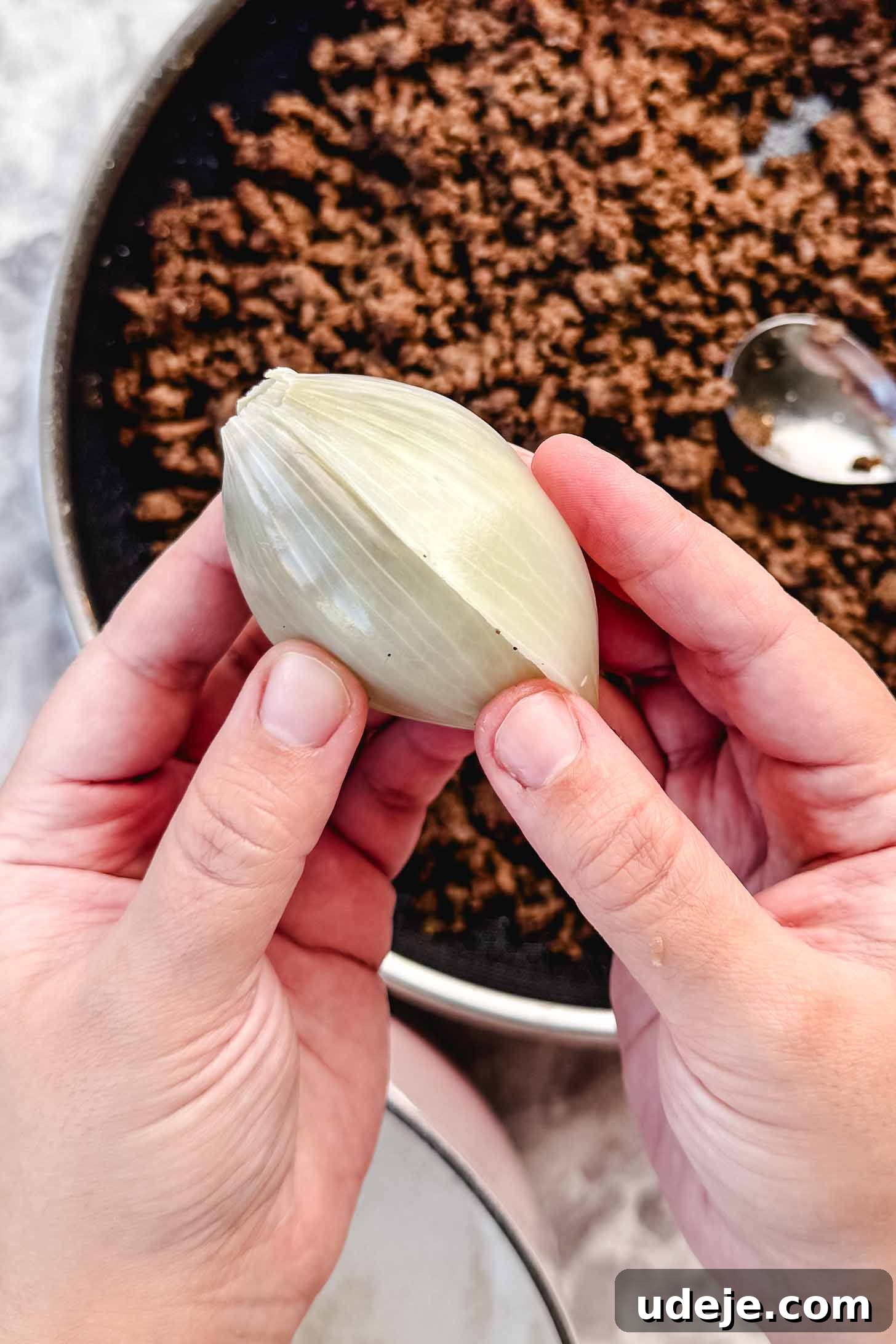 Hearty Beef-Stuffed Onions 11 A stuffed onion peel being held by two hands over a pan of cooked ground beef with a spoon in it on a marble background.