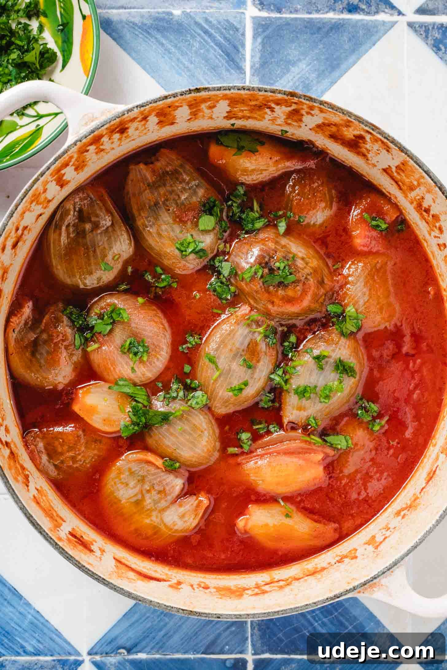 Hearty Beef-Stuffed Onions 14 White handled pot with stuffed onions in a tomato broth and chopped parsley on top on a blue and white tiled background with a bowl of chopped parsley at the side.