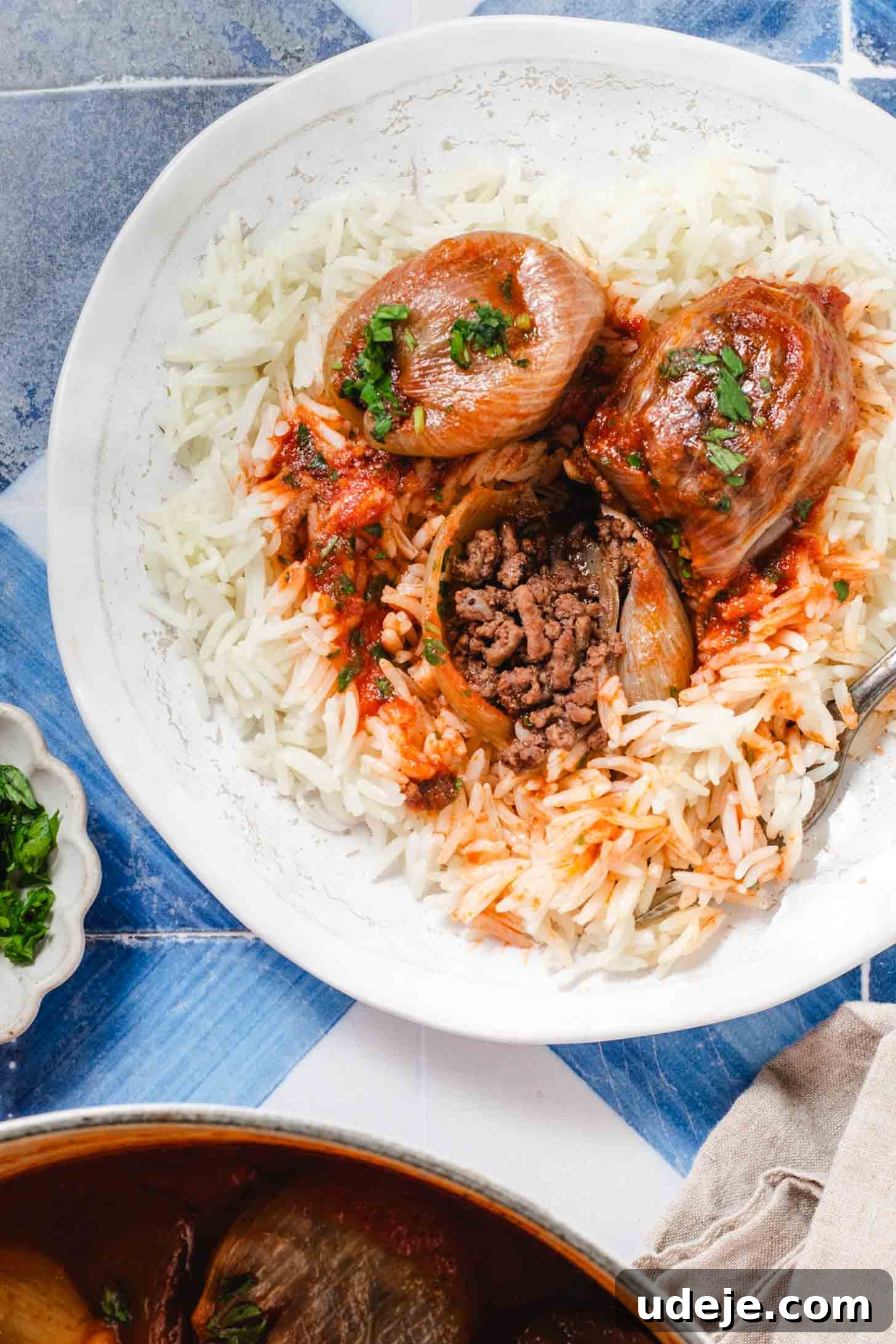 Hearty Beef-Stuffed Onions 15 A white plate with white rice and 3 stuffed onions with one cut open and a fork at the side on a blue and white tiled background, a small plate of parsley, a beige tea towel and white pot at the side.