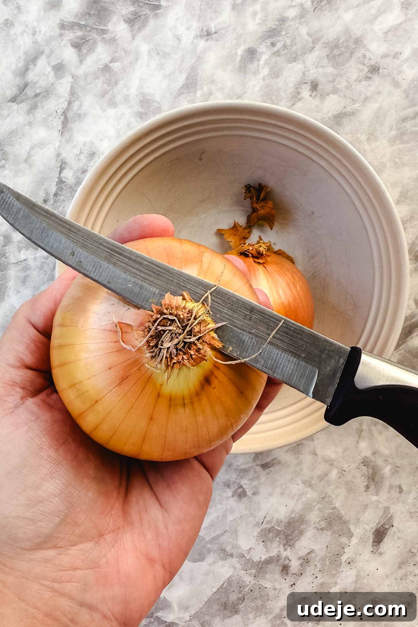 Hearty Beef-Stuffed Onions 4 Onion being held and the top being cut off with knife over a white bowl on a marble background.