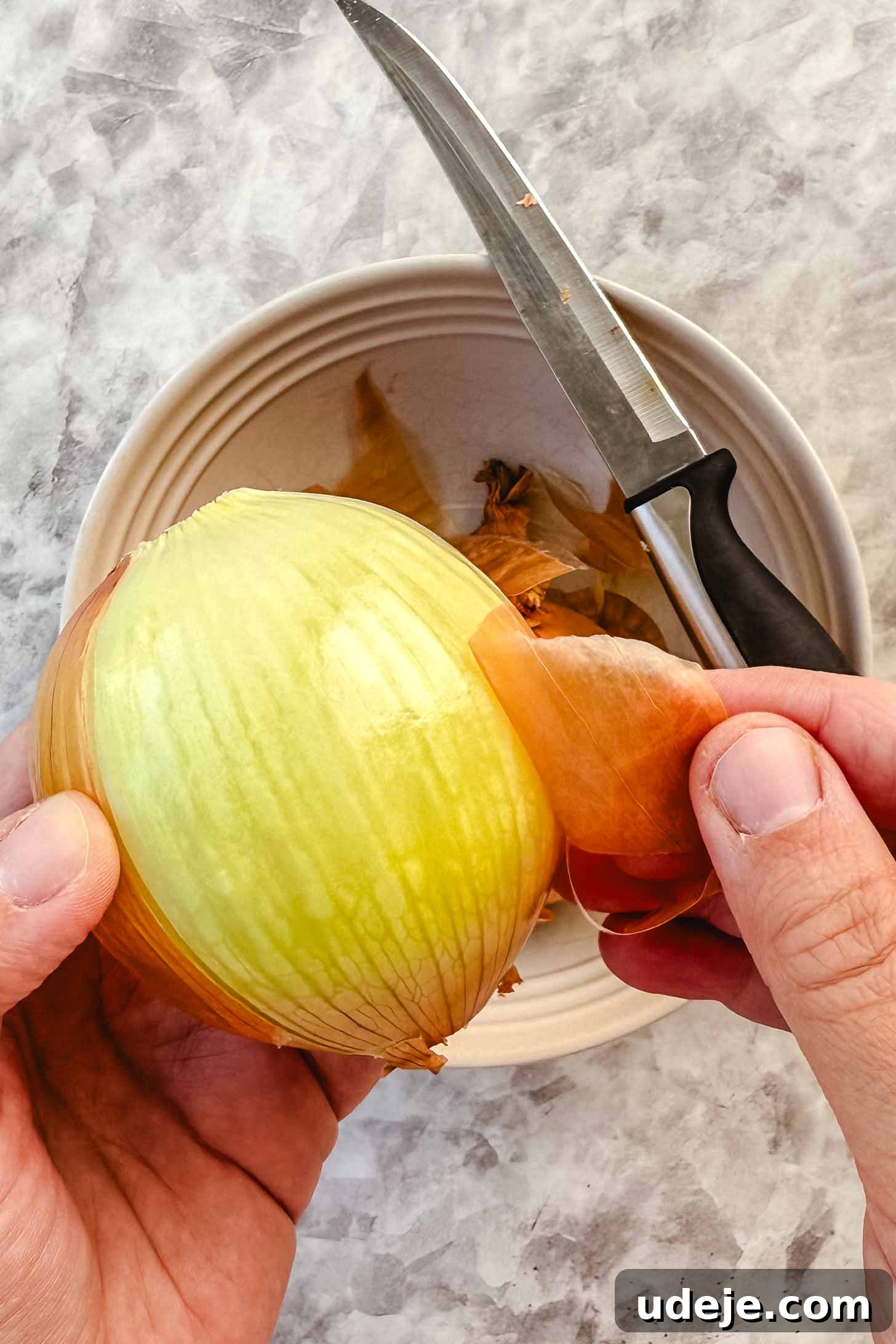 Hearty Beef-Stuffed Onions 6 Peeled onion being held over a white bowl with a knife with a black handle at the side on a marble background.