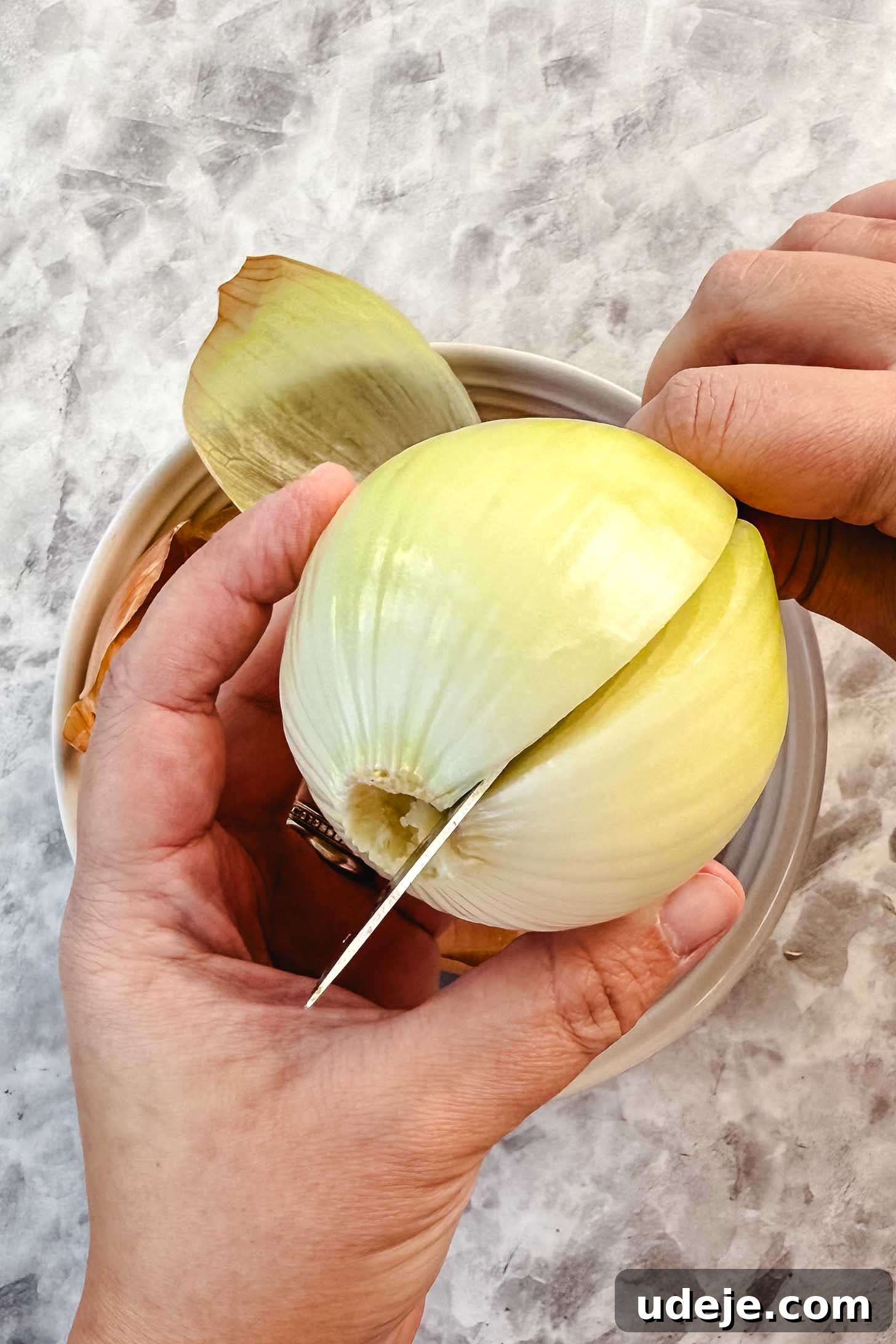 Hearty Beef-Stuffed Onions 7 Peeled onion being held with another hand cutting it in half over a white bowl with peels over a marble background.