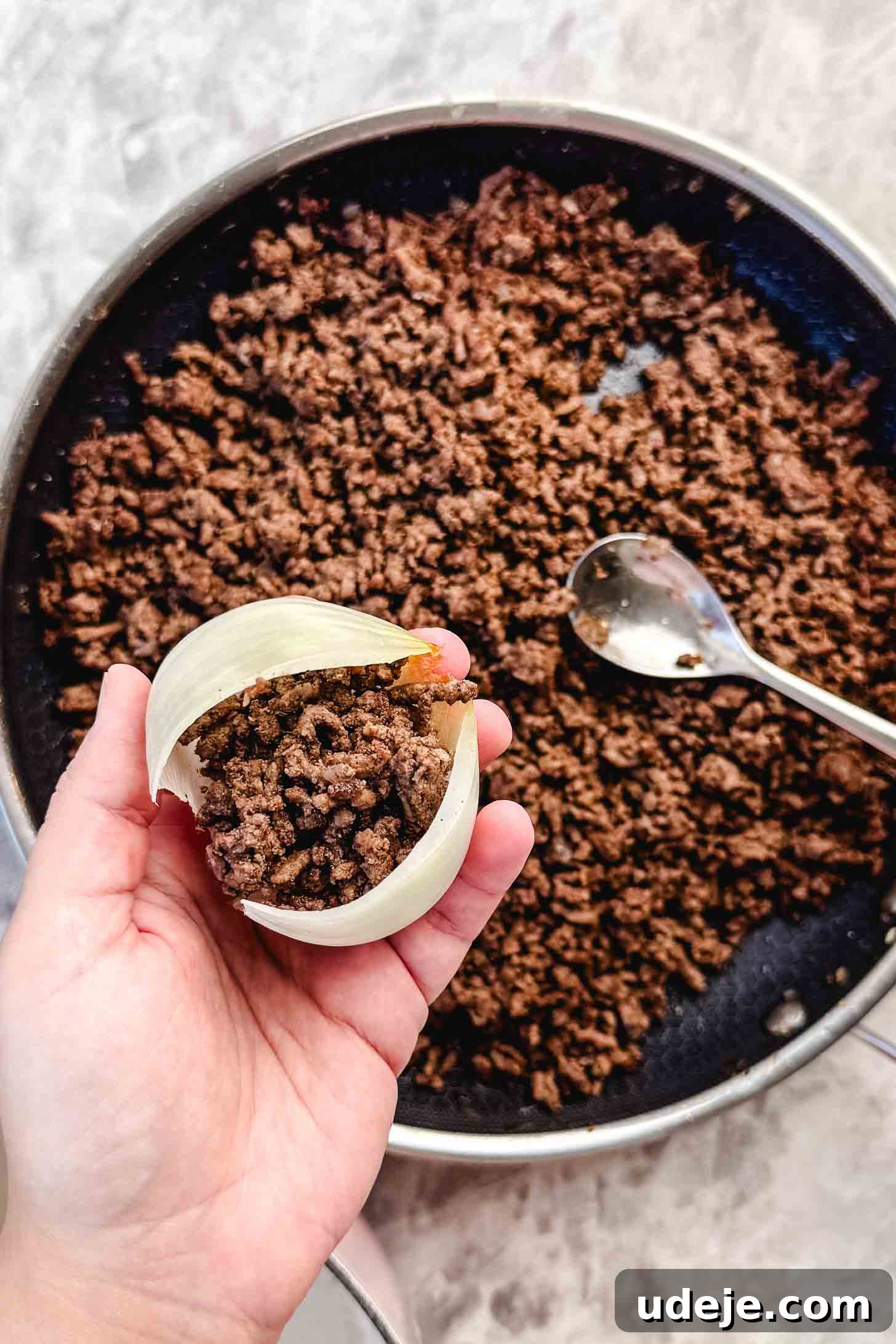 Hearty Beef-Stuffed Onions 10 Layer of onion being held by a hand with a cooked ground beef mixture inside it over a pan with cooked ground beef and a spoon on a marble background.