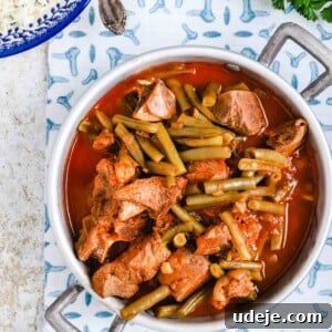Effortless Green Bean Stew Instant Pot and Stovetop 15 green bean stew with veal in a silver handled pot on a blue patterned tablecloth with rice in a blue plate and green onions and parsley on a light coloured plate on a beige marble background