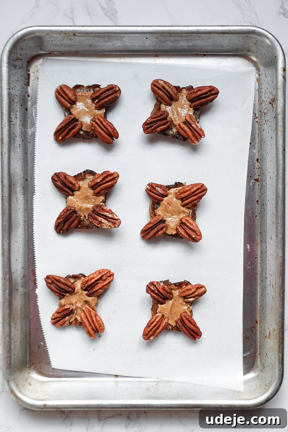 Easy 4-Ingredient Healthy Chocolate Turtles 6 Medjool dates on a parchment-lined tray, each topped with a dollop of peanut butter and meticulously placed pecan halves, forming the base of healthy chocolate turtles before coating.