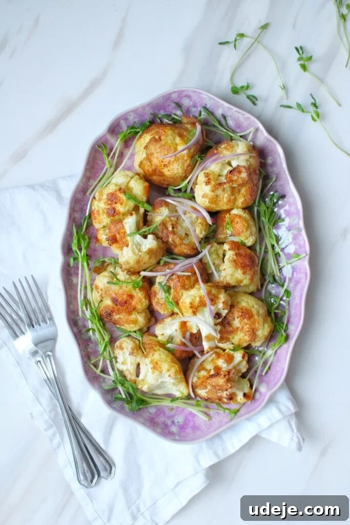 Crispy Cauliflower Bites 2 A pink oval plate with roasted cauliflower florets garnished with fresh microgreens, placed on a white napkin with two forks beside it on a light marble surface.