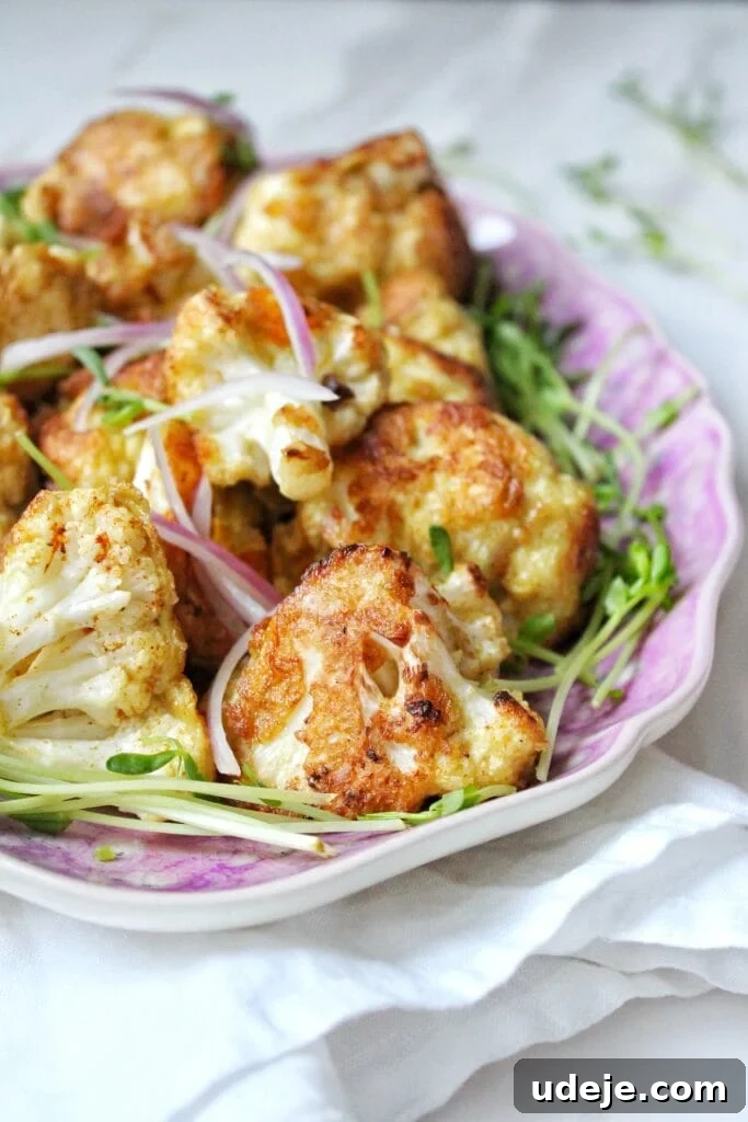 Crispy Cauliflower Bites 3 A close-up of roasted cauliflower florets garnished with thinly sliced red onions and fresh green sprouts, served on a purple-edged plate set on a white cloth.