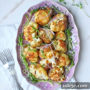 A pink oval plate with roasted cauliflower florets garnished with fresh microgreens, set on a white napkin with two forks beside it on a marble surface.