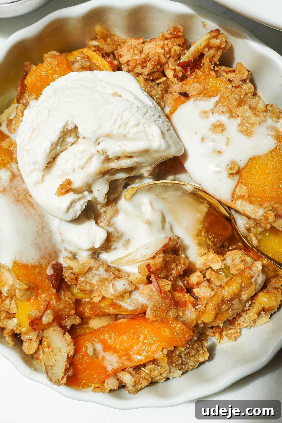 Wholesome Peach Crumble 4 A spoon delicately scooping a portion of warm peach crisp, revealing the bubbly fruit filling beneath the inviting nutty oat topping, with melting vegan ice cream on top.
