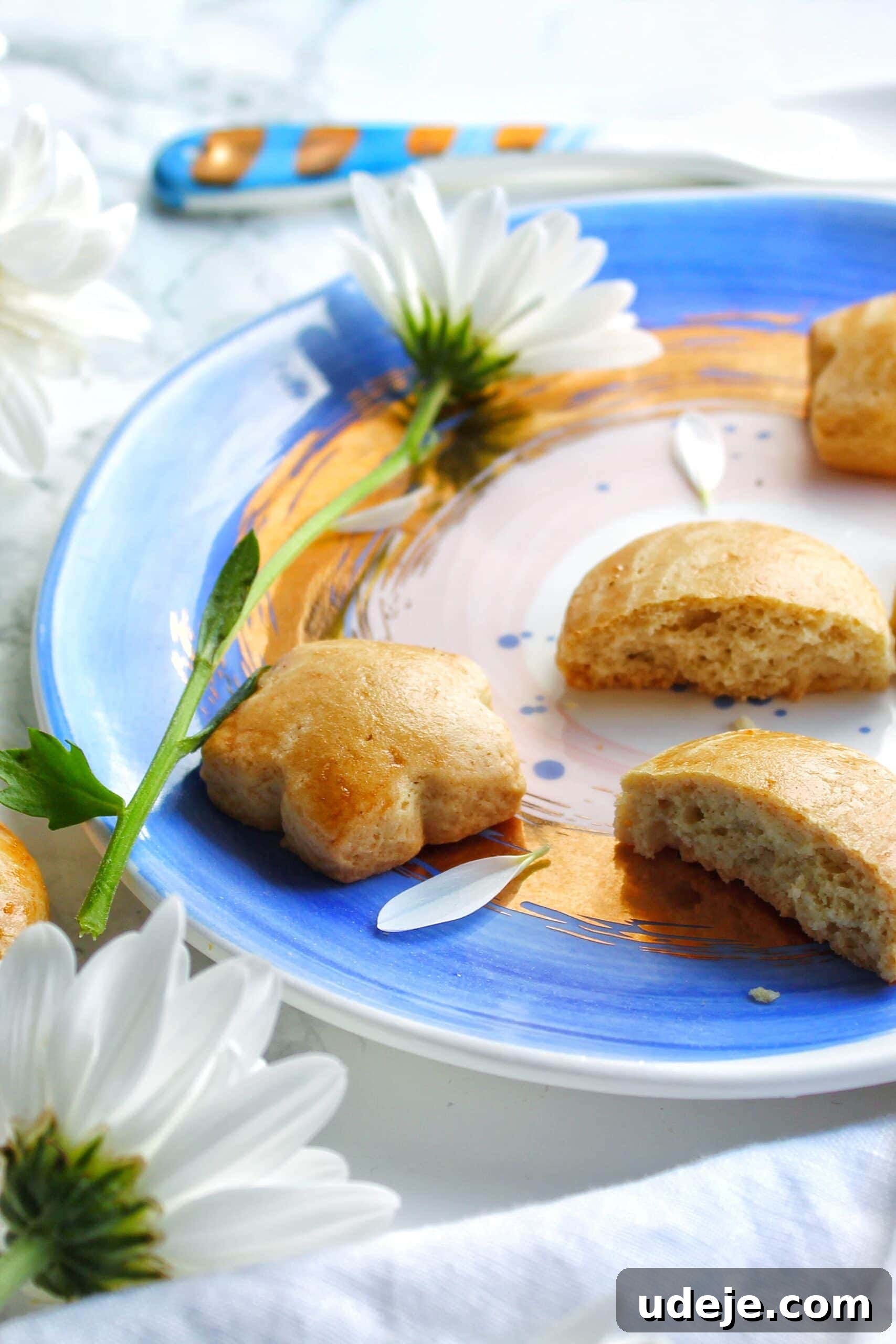 Traditional Iraqi Soda Biscuits 3 A blue and white plate with partially eaten Iraqi Soda Cookies, surrounded by white daisies, on a light surface with a butter knife in the background, showcasing their irresistible appeal.