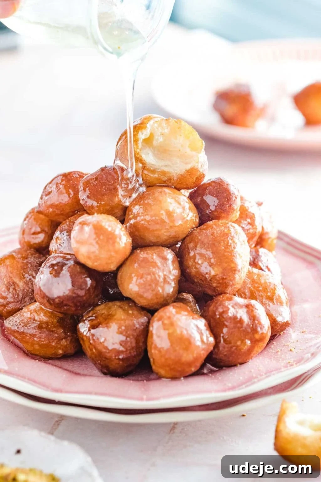 Crunchy Middle Eastern Luqaimat and Awamat Fritters 2 Golden Luqaimat (fried dough balls) are elegantly stacked on a pink plate, being generously drizzled with glistening sugar syrup from a clear jug. This image beautifully captures the essence of this beloved Middle Eastern dessert, perfect for Ramadan celebrations.