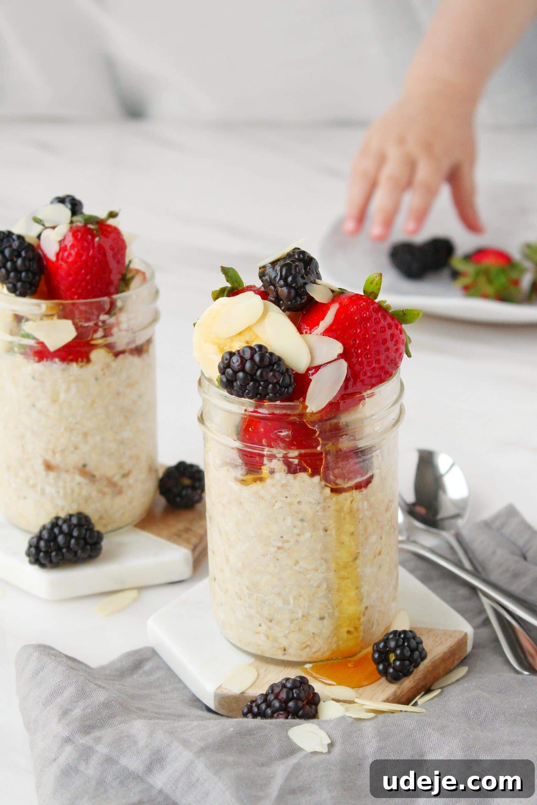 Effortless Morning Oats 3 Two aesthetically arranged glass jars of overnight oats, brimming with oats and topped with a vibrant medley of fresh strawberries, dark blackberries, crisp almond slices, and a glistening drizzle of honey. In the soft-focus background, a child’s hand gently reaches for berries on a plate, adding a touch of wholesome family life. Two silver spoons rest gracefully on a textured gray napkin beside the jars, completing the inviting breakfast scene.