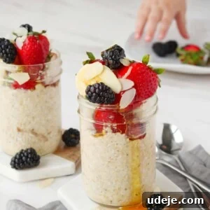 Two jars of overnight oats topped with strawberries, blackberries, almond slices, and honey. A child’s hand reaches for berries on a plate in the background. Two spoons rest on a gray napkin beside the jars.