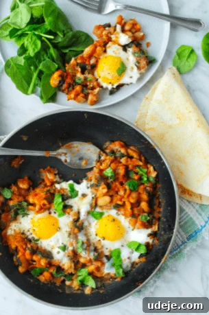 Savory Skillet Brunch: Eggs and Beans 2 A black skillet filled with a vibrant mixture of cooked beans, spinach, and sunny-side-up eggs. A golden tortilla rests on the side, and a white plate nearby holds a serving of the eggs and beans with fresh spinach leaves, highlighting the dish's appetizing presentation.