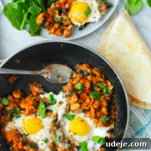 Black skillet with cooked beans and sunny side up eggs with a tortilla at the side and a white plate with the eggs and beans and spinach leaves beside it.