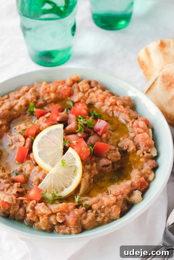 Authentic Egyptian Ful Medames 2 A bowl of Ful Medames fava bean dip garnished with chopped tomatoes, parsley, a drizzle of olive oil, and two lemon slices, served with pita bread on the side and green glasses in the background.