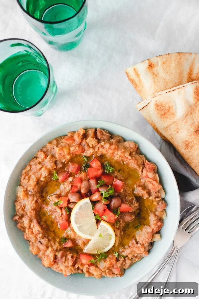 Authentic Egyptian Ful Medames 3 A bowl of creamy Egyptian beans, or Ful Medames, topped with diced tomatoes, herbs, and lemon slices sits next to pita bread and two green glasses of water on a white tablecloth. Two forks are placed beside the bowl.
