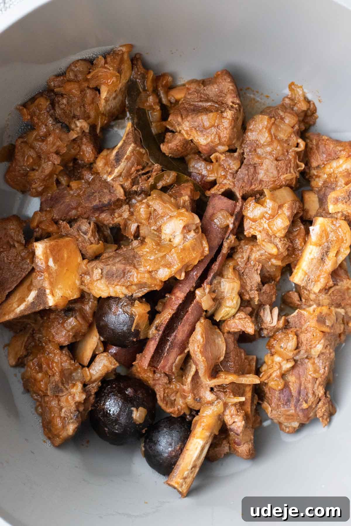 Chef's Signature Lamb Kabsa 11 Cooked lamb meat pieces with bones, onions, whole spices like cinnamon sticks and bay leaves, and black dried fruits or seeds in a gray pot—a savory dish reminiscent of lamb kabsa, aromatic and well-seasoned.