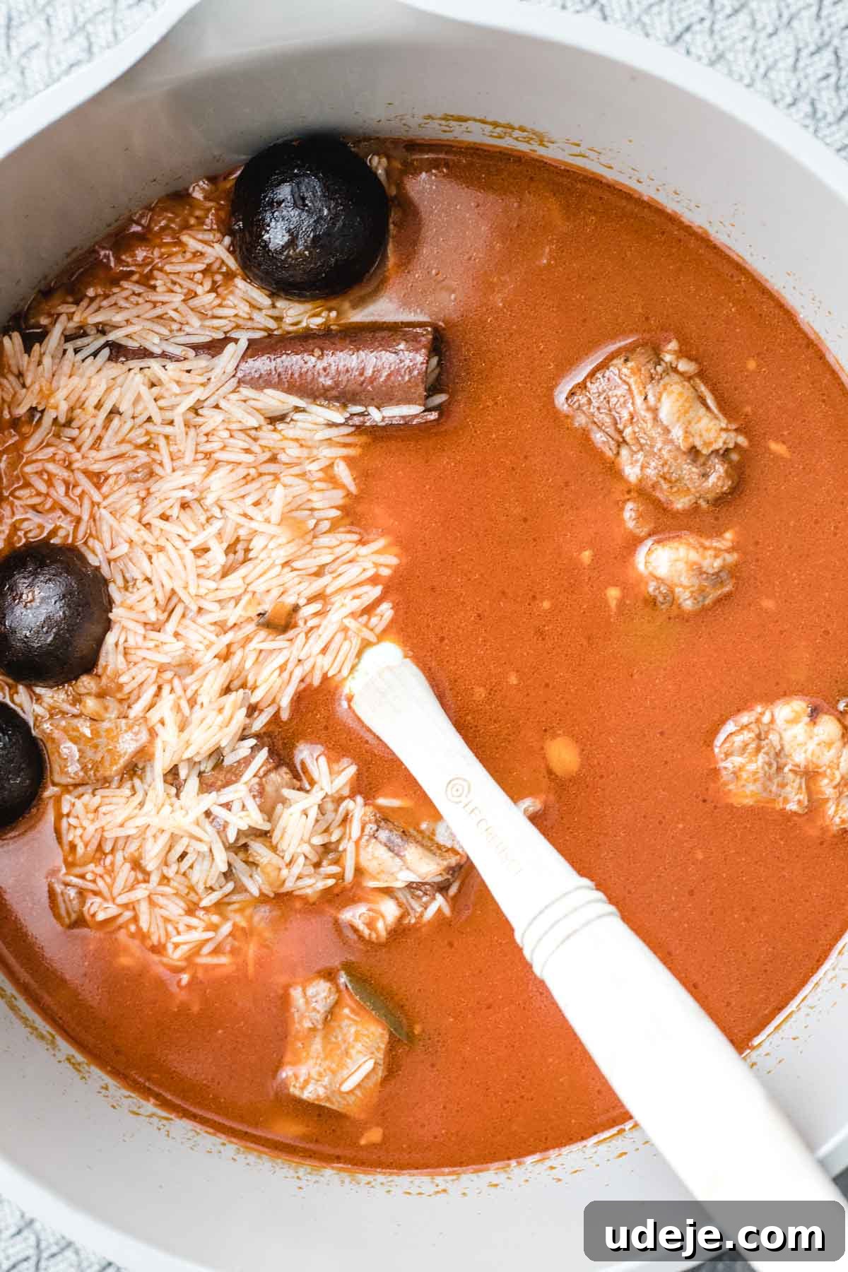 Chef's Signature Lamb Kabsa 12 A white bowl contains reddish-brown lamb kabsa broth, raw white rice, chunks of meat, whole dried limes, and a cinnamon stick, with a white spoon resting inside.