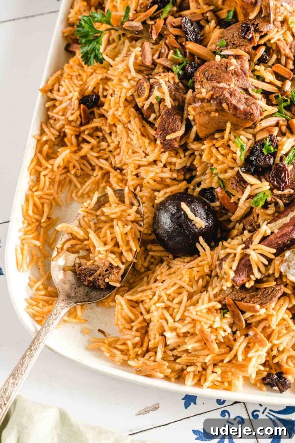 Chef's Signature Lamb Kabsa 14 Close up shot of lamb kabsa served in a dish and garnished with parsley, almonds and raisins. The dish is on a tiled surface and has a spoon inside.