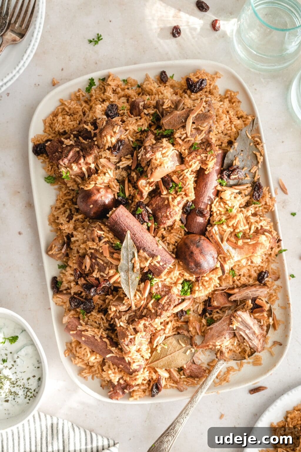 Chef's Signature Lamb Kabsa 3 Lamb and rice served on a white platter with a silver spoon, with a glass of water and cup of yogurt nearby.