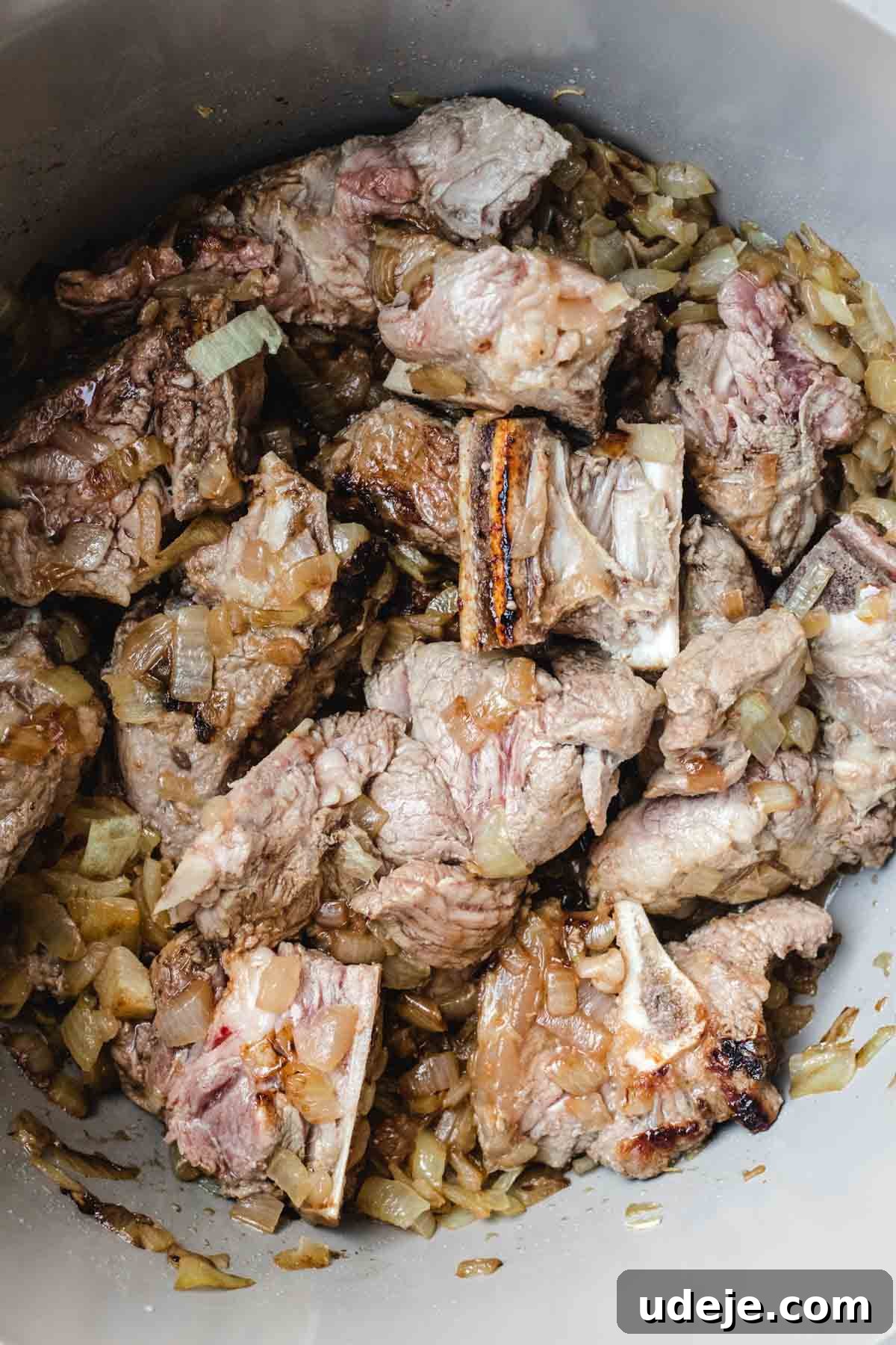 Chef's Signature Lamb Kabsa 6 Chunks of browned lamb with bones are cooking with sautéed chopped onions in a pot, reminiscent of the rich flavors found in lamb kabsa. The meat is seared and mixed with translucent, slightly caramelized onions.