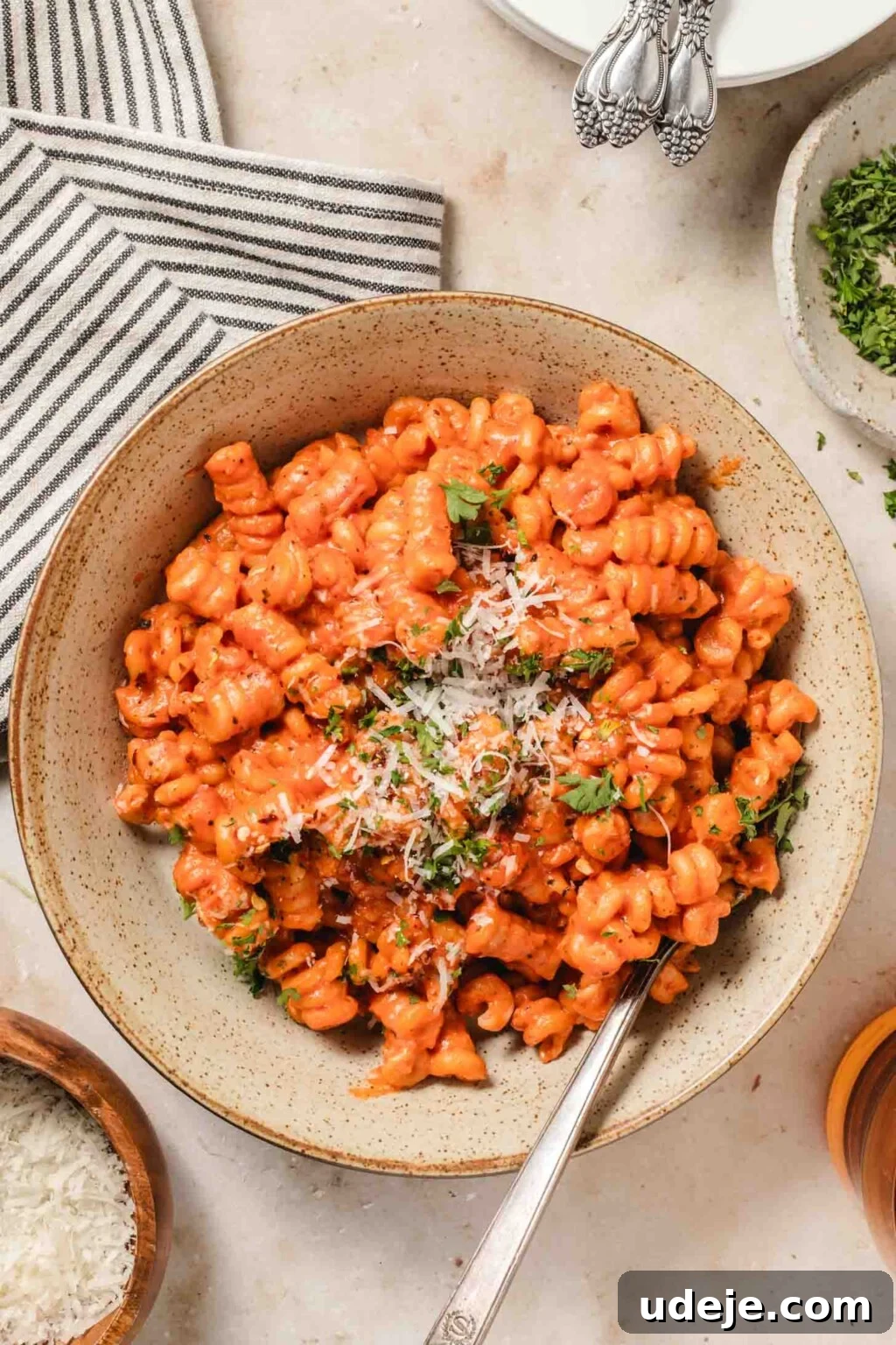 30-Minute One-Pot Creamy Pink Pasta 2 creamy pink sauce pasta in fusili shape is plated in a brown speckled pasta bowl, garnished with parmesan and chopped parsley. It is on a table with a striped napkin, a small parmesan dish, and some parsley in a small dish.