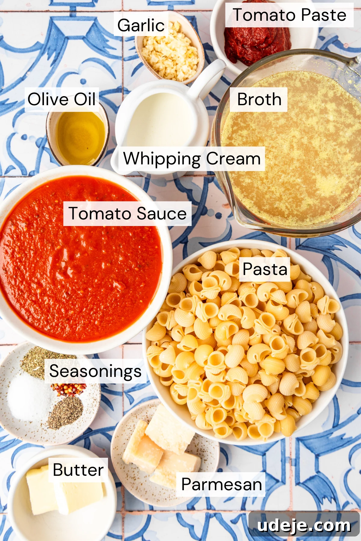 30-Minute One-Pot Creamy Pink Pasta 4 Ingredient shot with a small white bowl with tomato paste, a small bowl with minced garlic, a measuring cup with broth, a small white pitcher with whipping cream, a small bowl with olive oil, a white bowl with tomato sauce, a white bowl with dried pasta, a small plate with seasonings, a small plate with chunks of parmesan and a small white bowl with butter on a blue and white tiled background.