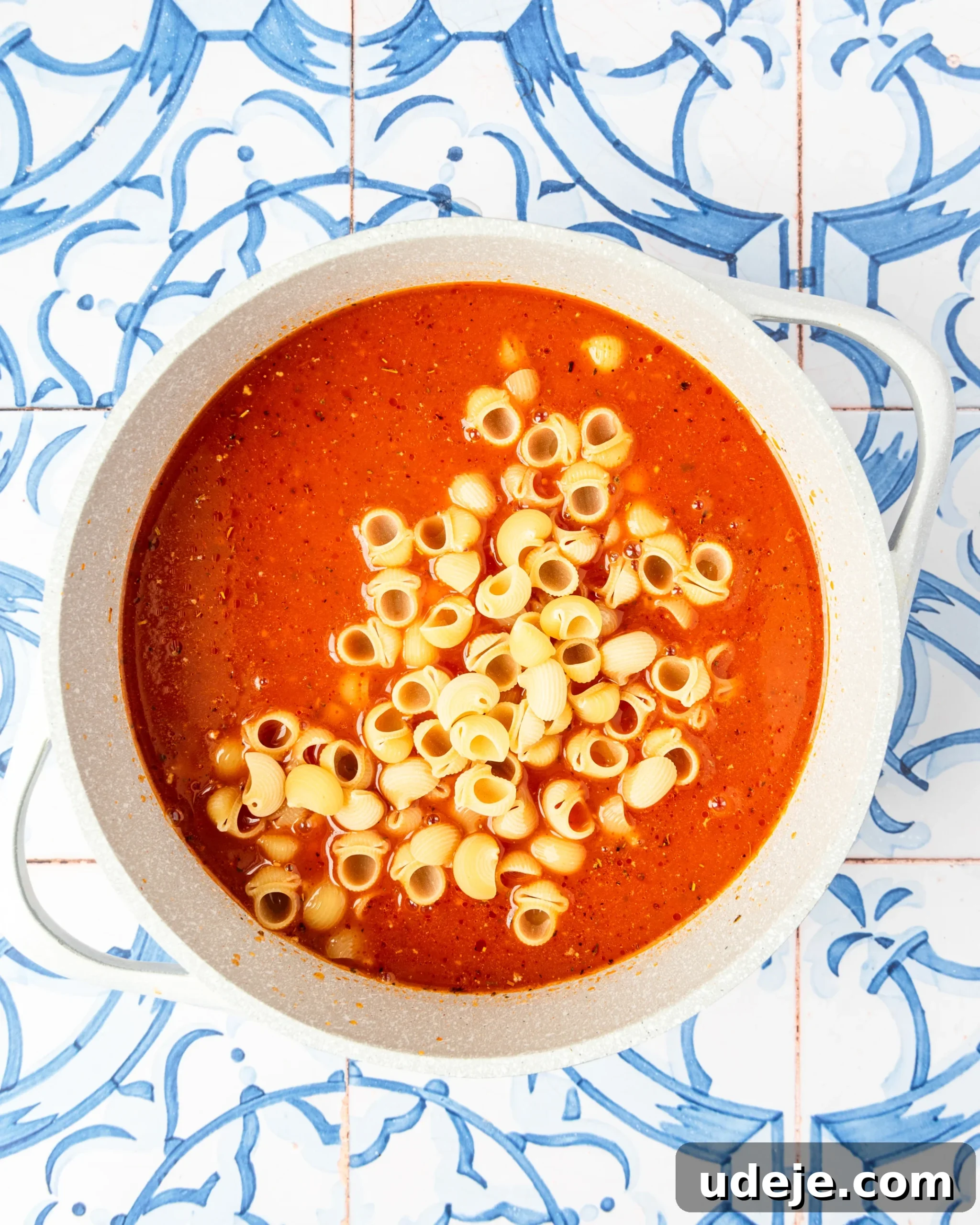 30-Minute One-Pot Creamy Pink Pasta 9 White pot with red liquid and seasonings along with shell pasta on a blue and white tiled background.