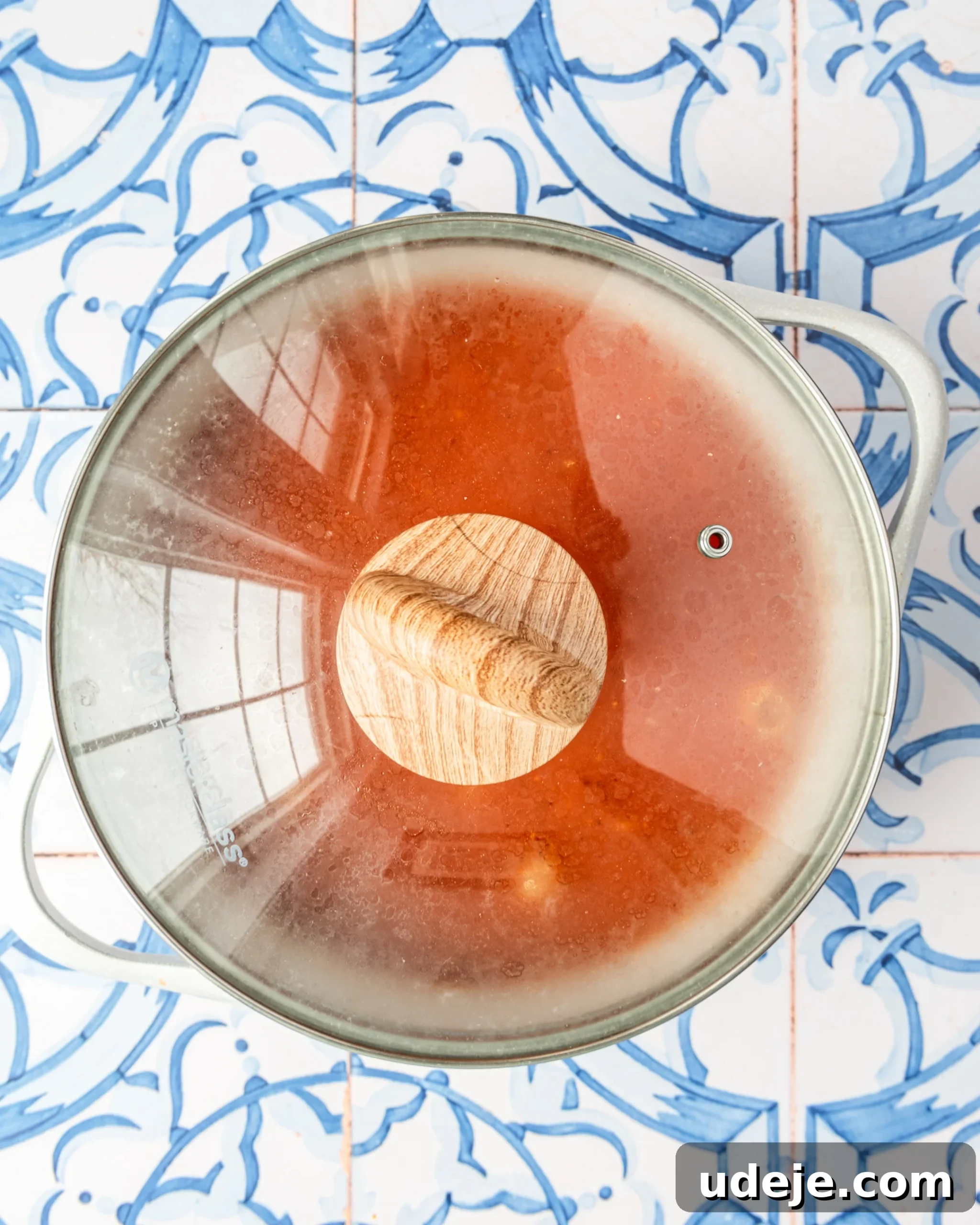 30-Minute One-Pot Creamy Pink Pasta 10 White pot with red liquid and pasta mixed in covered with a glass lid with a wooden handle on top on a blue and white tiled background.