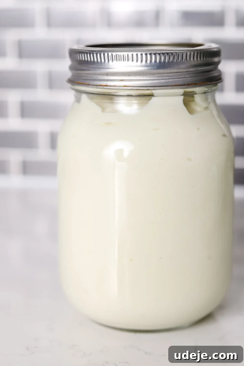 Whip Up Fluffy Toum in 5 Minutes No Food Processor 12 A perfectly sealed glass mason jar, filled with creamy white Toum, stands on a clean white countertop against a backdrop of gray and white tiled backsplash, ready for storage.
